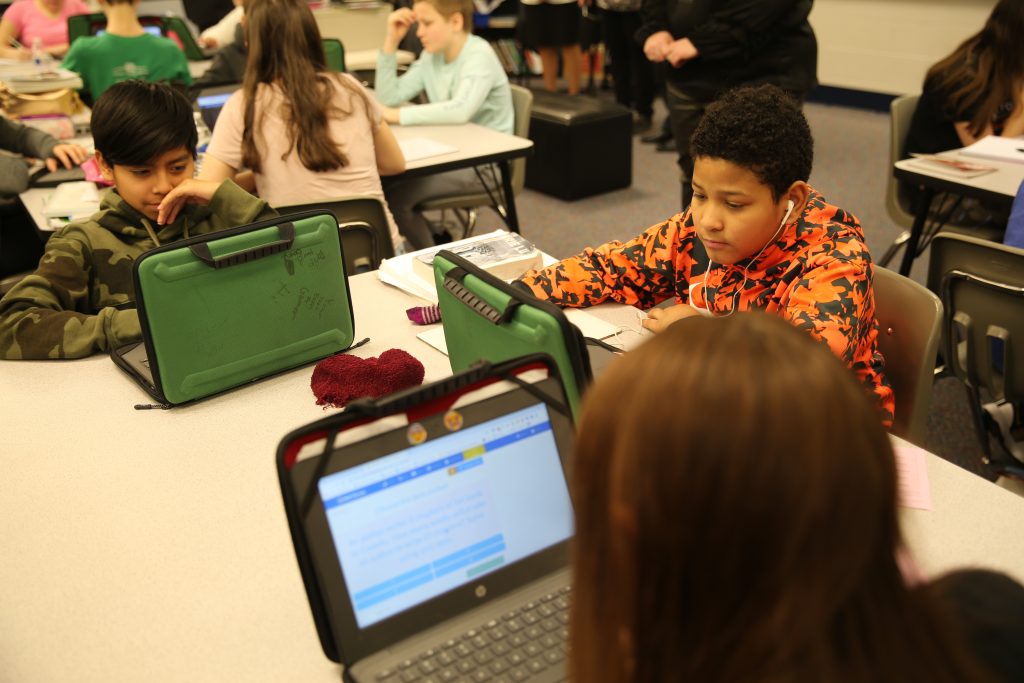 Two children sitting at a table in a classroom, each engrossed in their own laptop, surrounded by other students who are